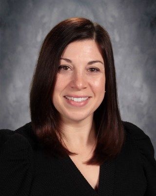 Woman with brown hair smiles, wearing a black top against a blurred gray background.