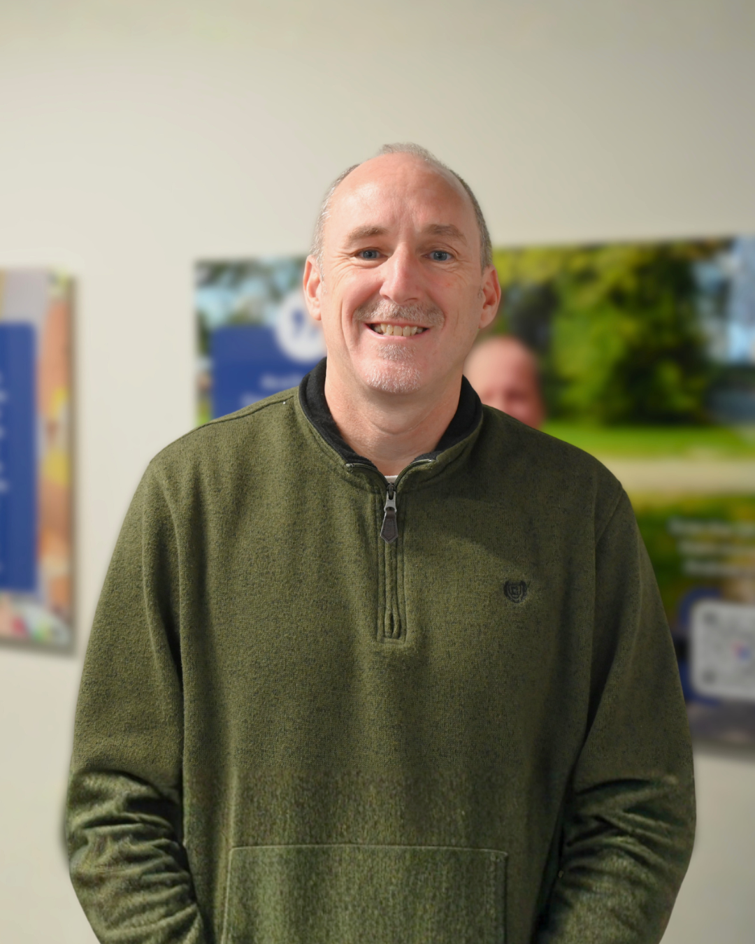 Man with light skin wearing a brown shirt and patterned tie smiling at the camera against a neutral backdrop.