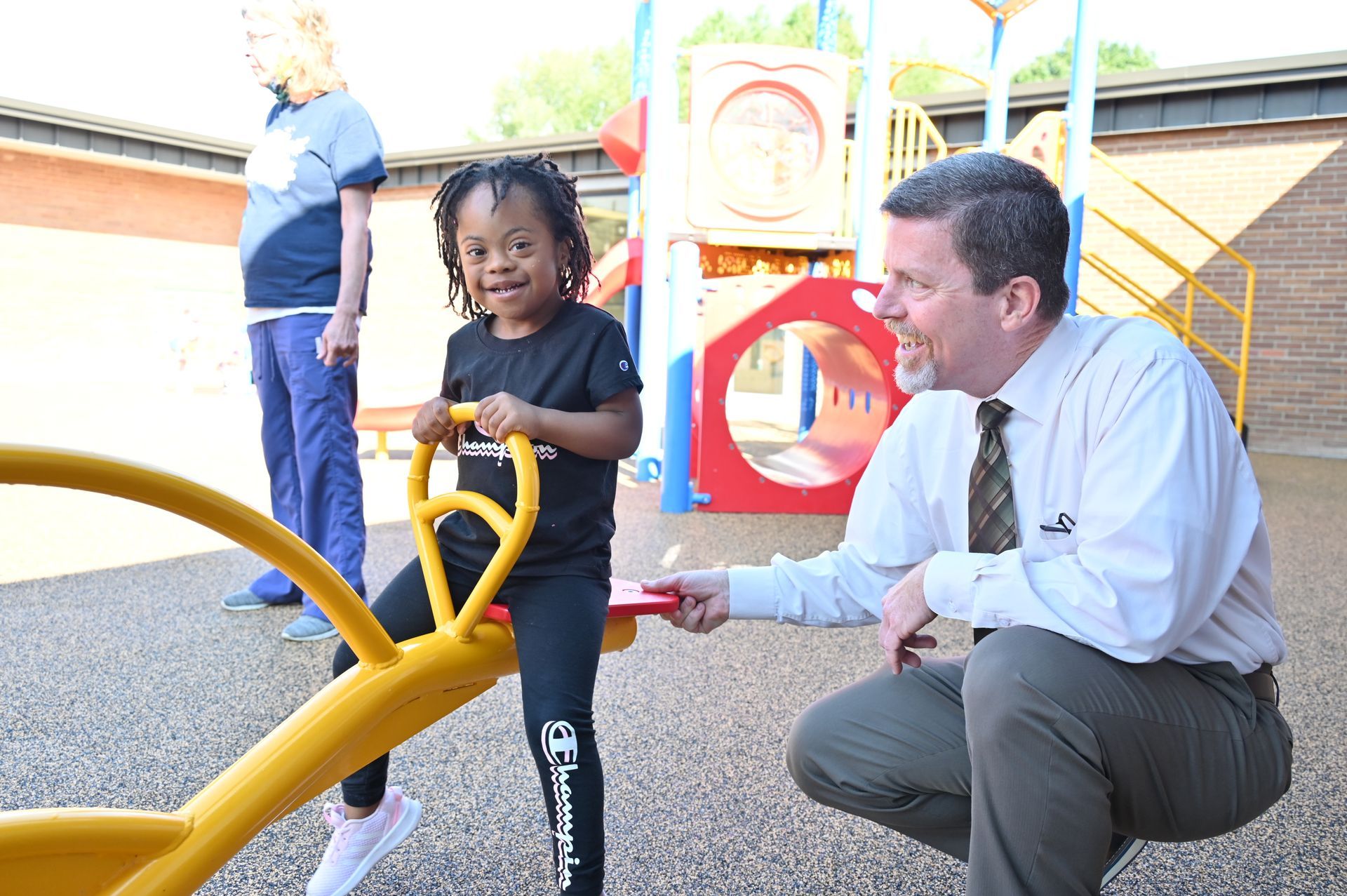 Child on a yellow seesaw at a playground smiles, being assisted by an adult male. Another person watches.
