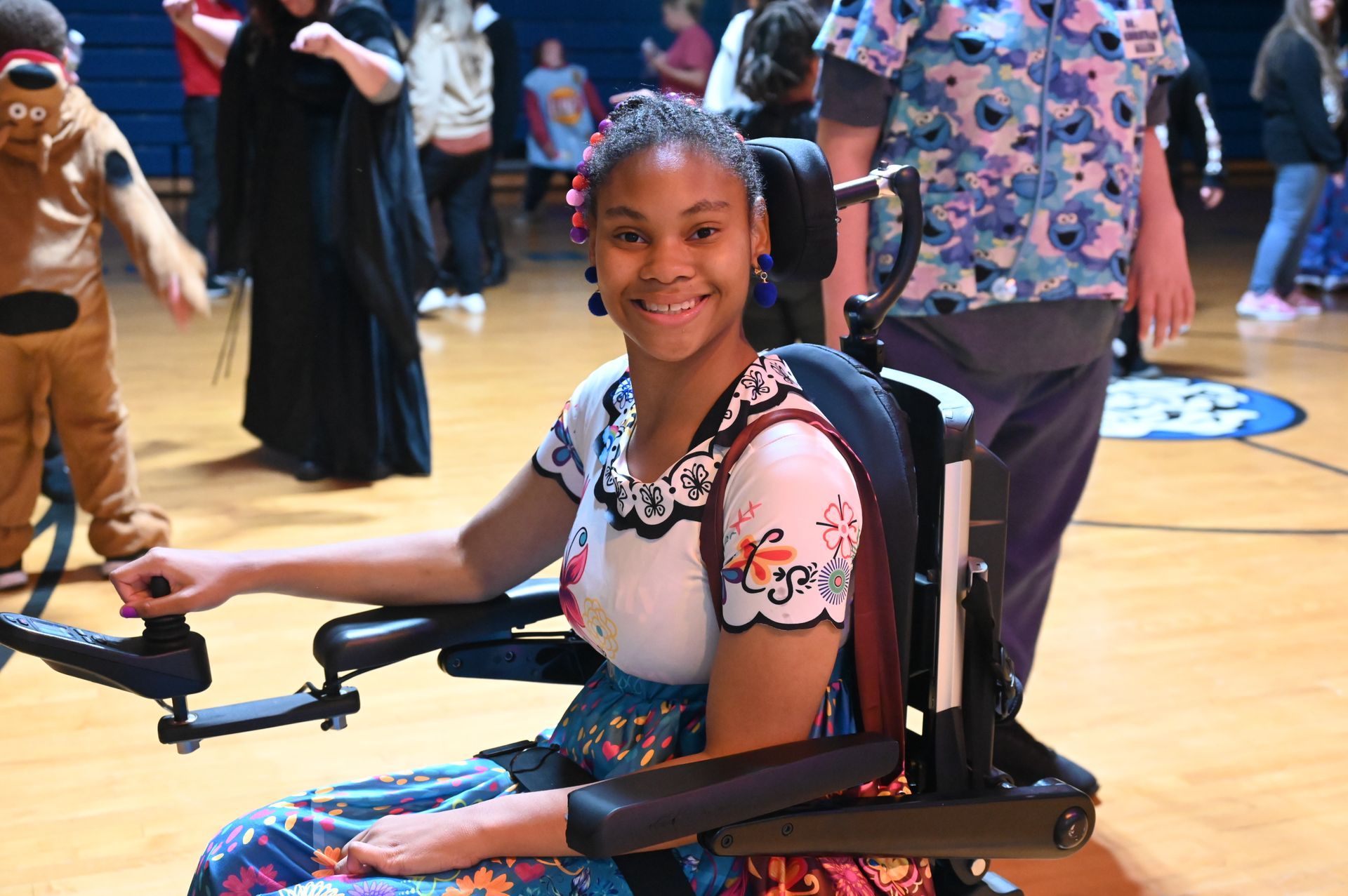 Smiling person in wheelchair, wearing colorful floral dress, in gym; people dance in the background.