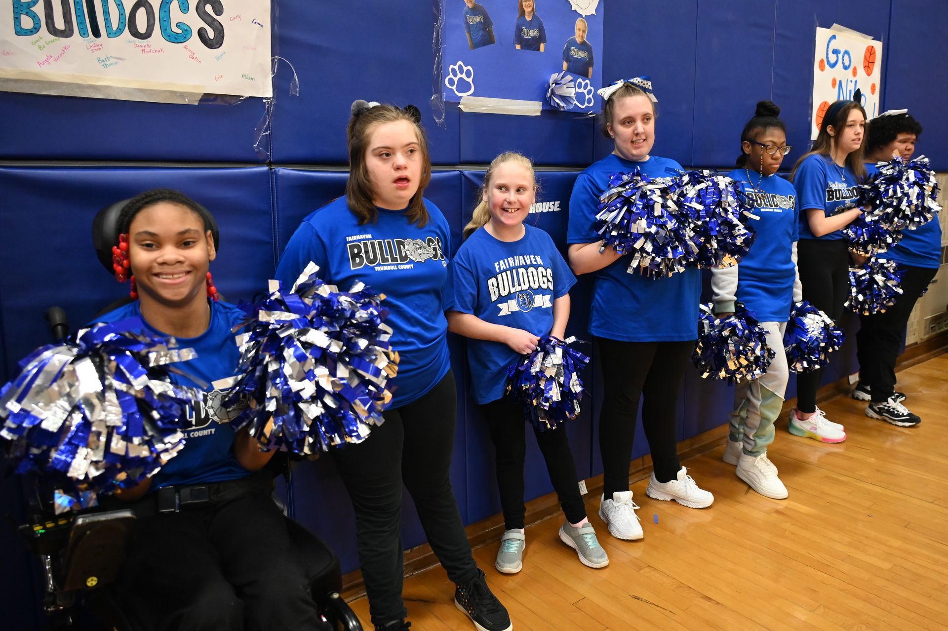Cheerleaders in blue, holding pom-poms, smiling. Gym setting, Bulldog sign in background.