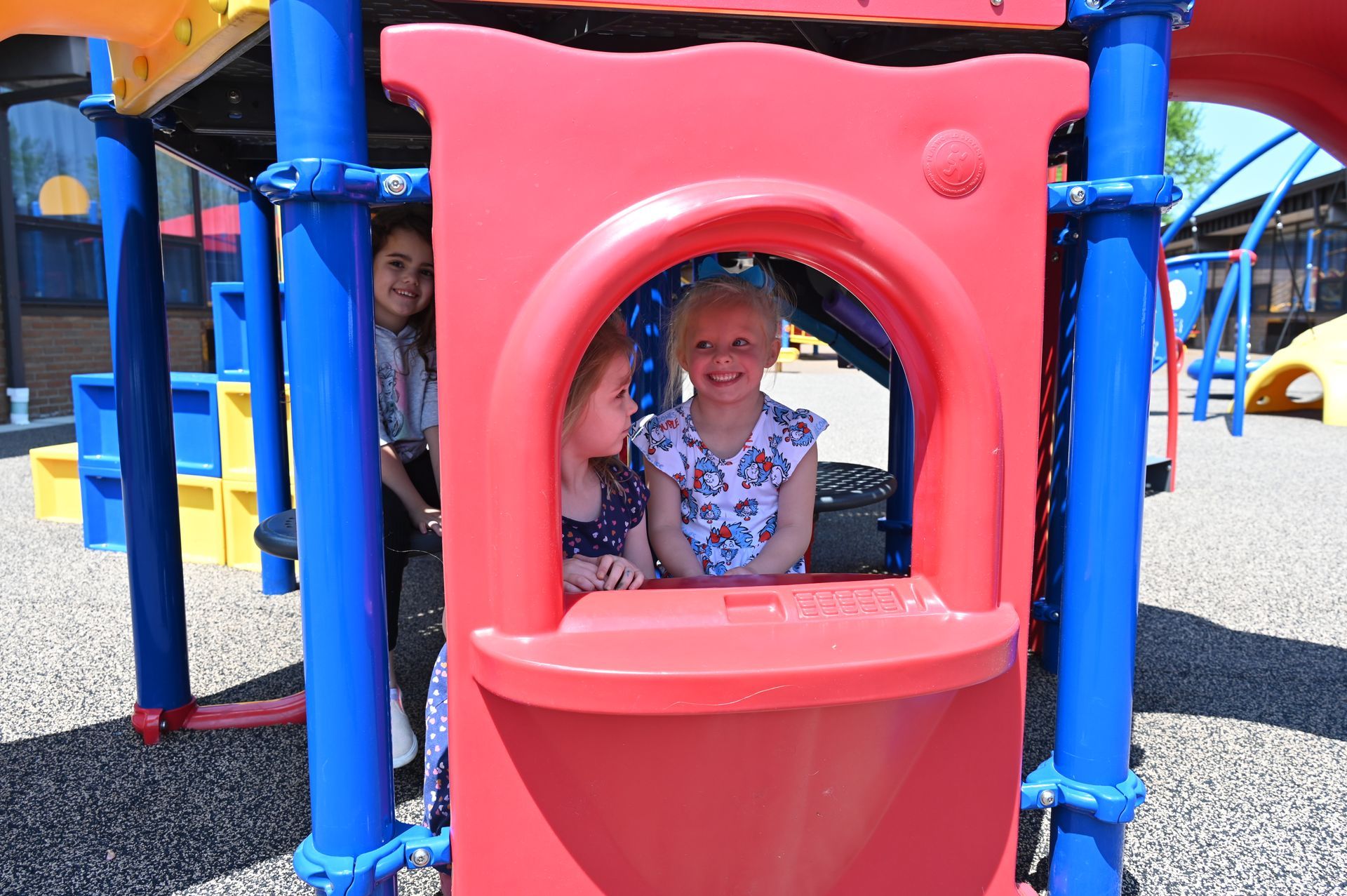 Three children smiling in a red play structure; one looking out, two peeking through the archway.