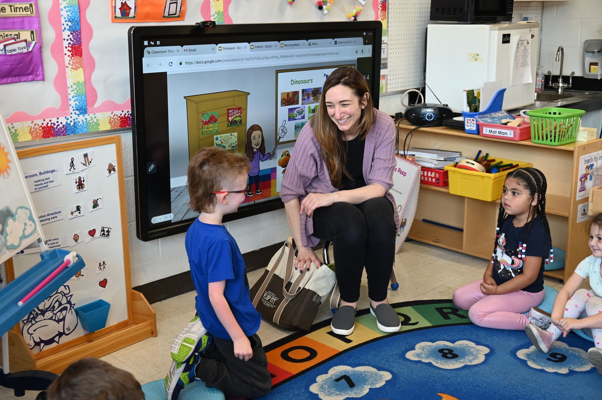 Teacher with children in a classroom, gathered on a rug. The teacher smiles, pointing at a screen.