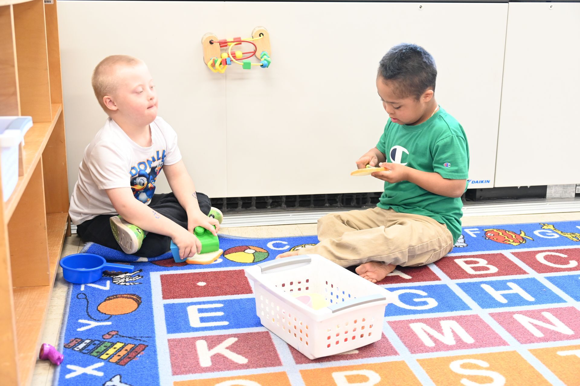 Two children sitting on a rug with toys, one holding a small item, the other looking on.
