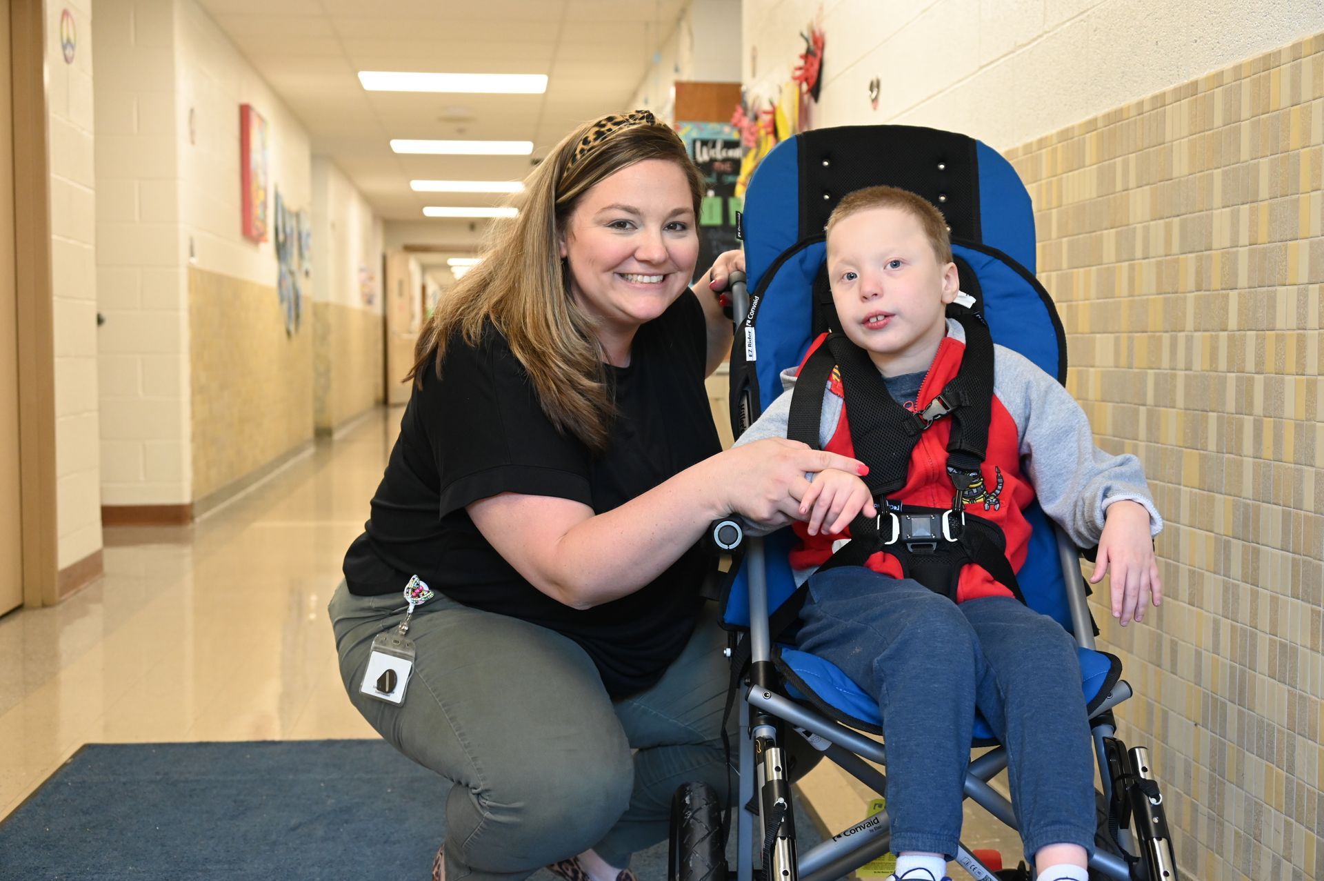 Woman and boy in wheelchair smile in hallway. The woman kneels, holding the boy's hand.