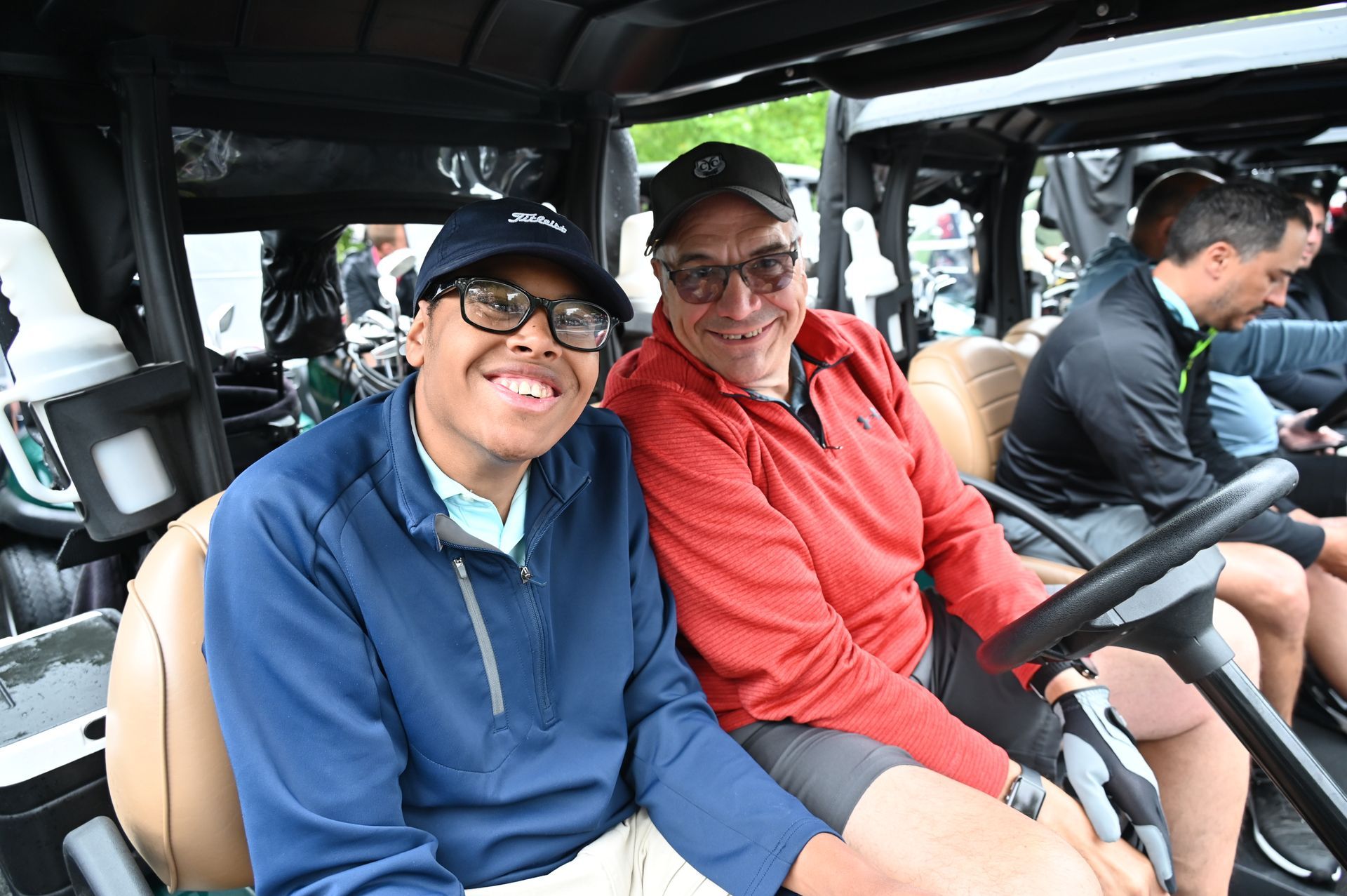 Two men smiling in a golf cart. One wears a blue shirt and glasses, the other a red jacket. 