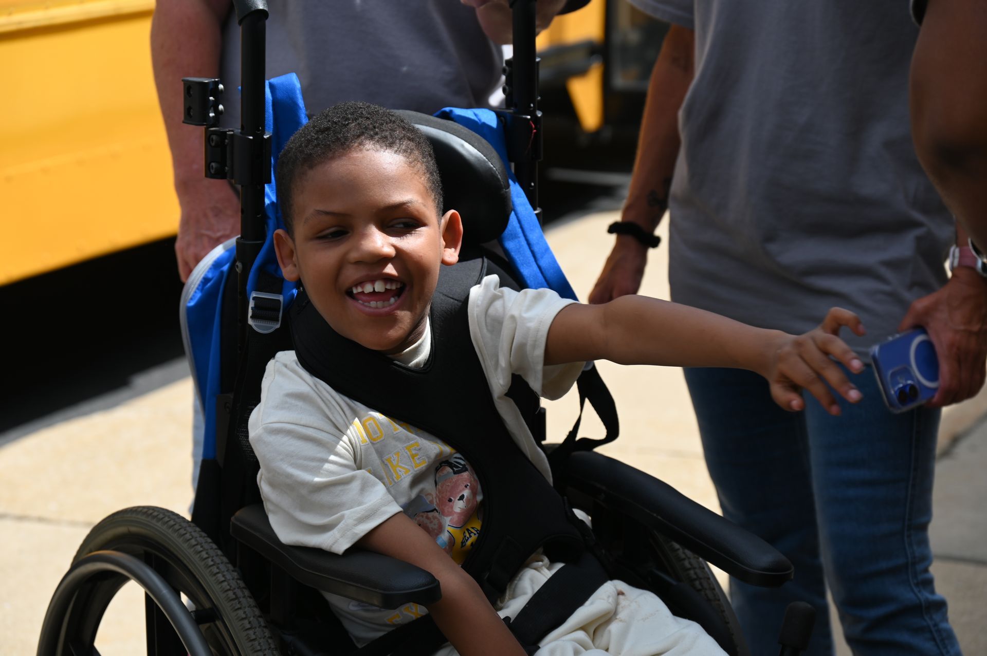Boy in wheelchair smiles and reaches out. Near a yellow school bus.