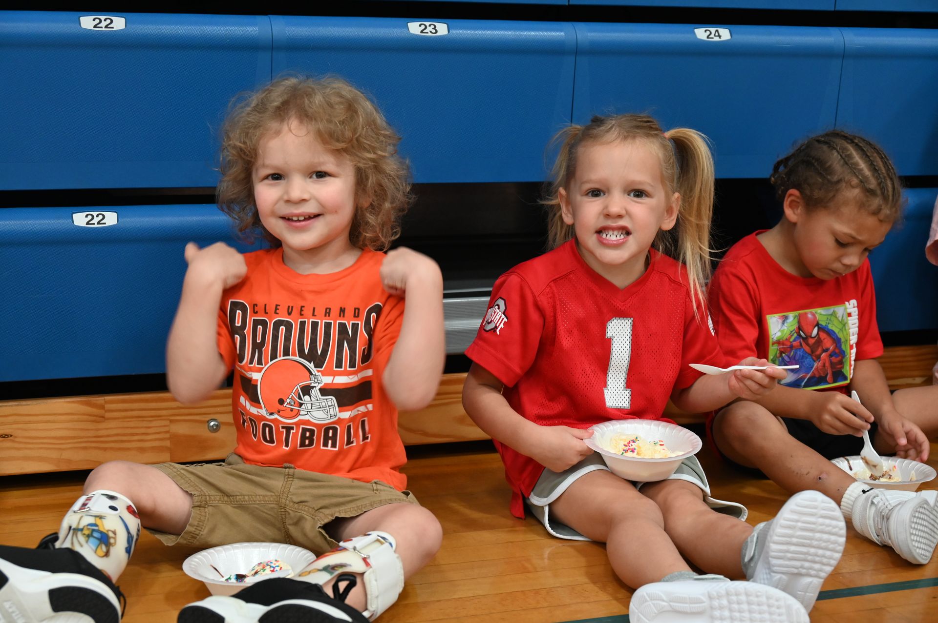 Three children seated on a floor smiling with bowls of food in their laps; one in orange Browns shirt.
