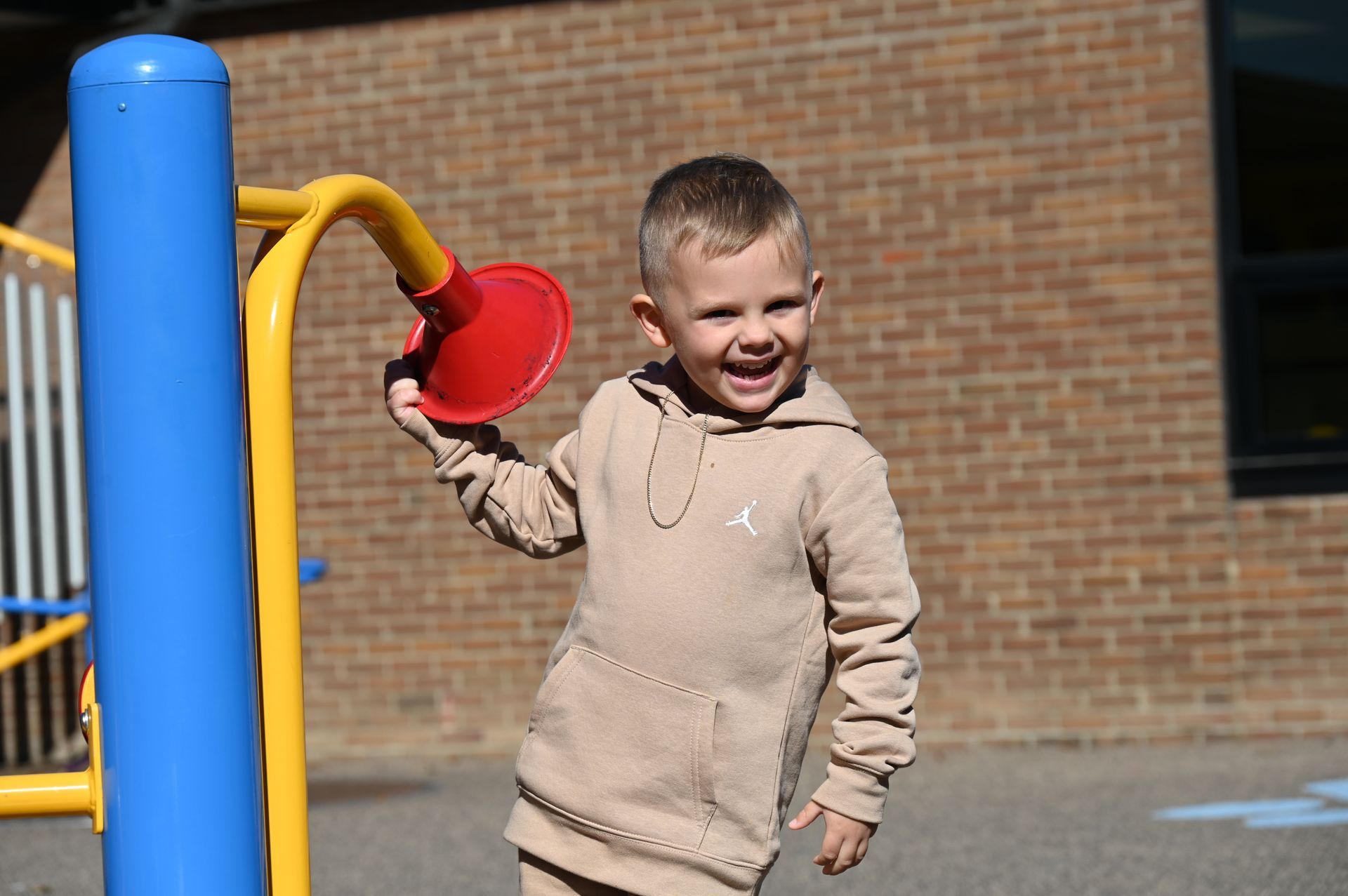 Boy smiling while holding red object on playground. Wearing tan hoodie and pants.