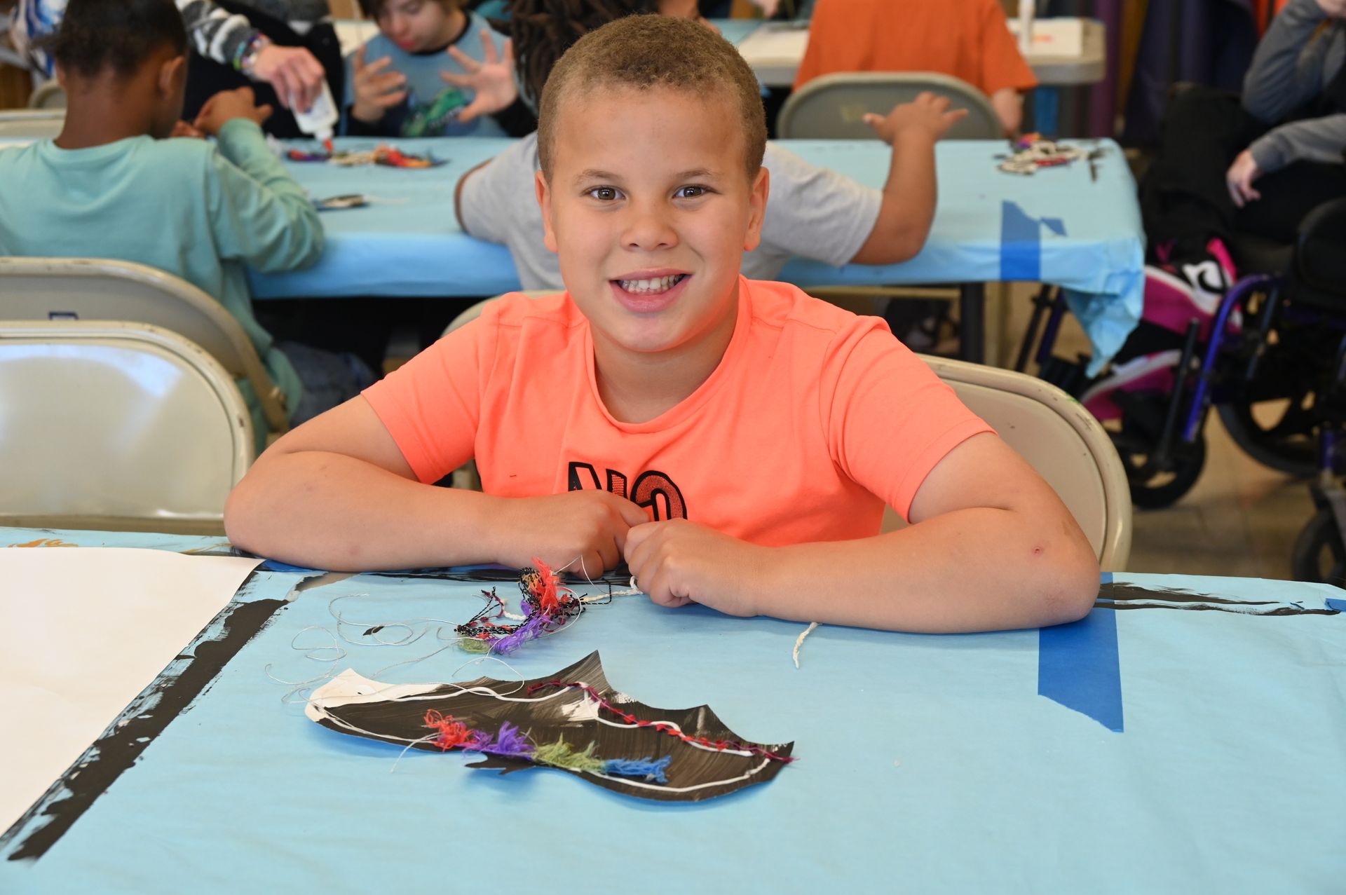 Boy smiling at a craft table, holding bat-shaped artwork. Other children and tables in background.