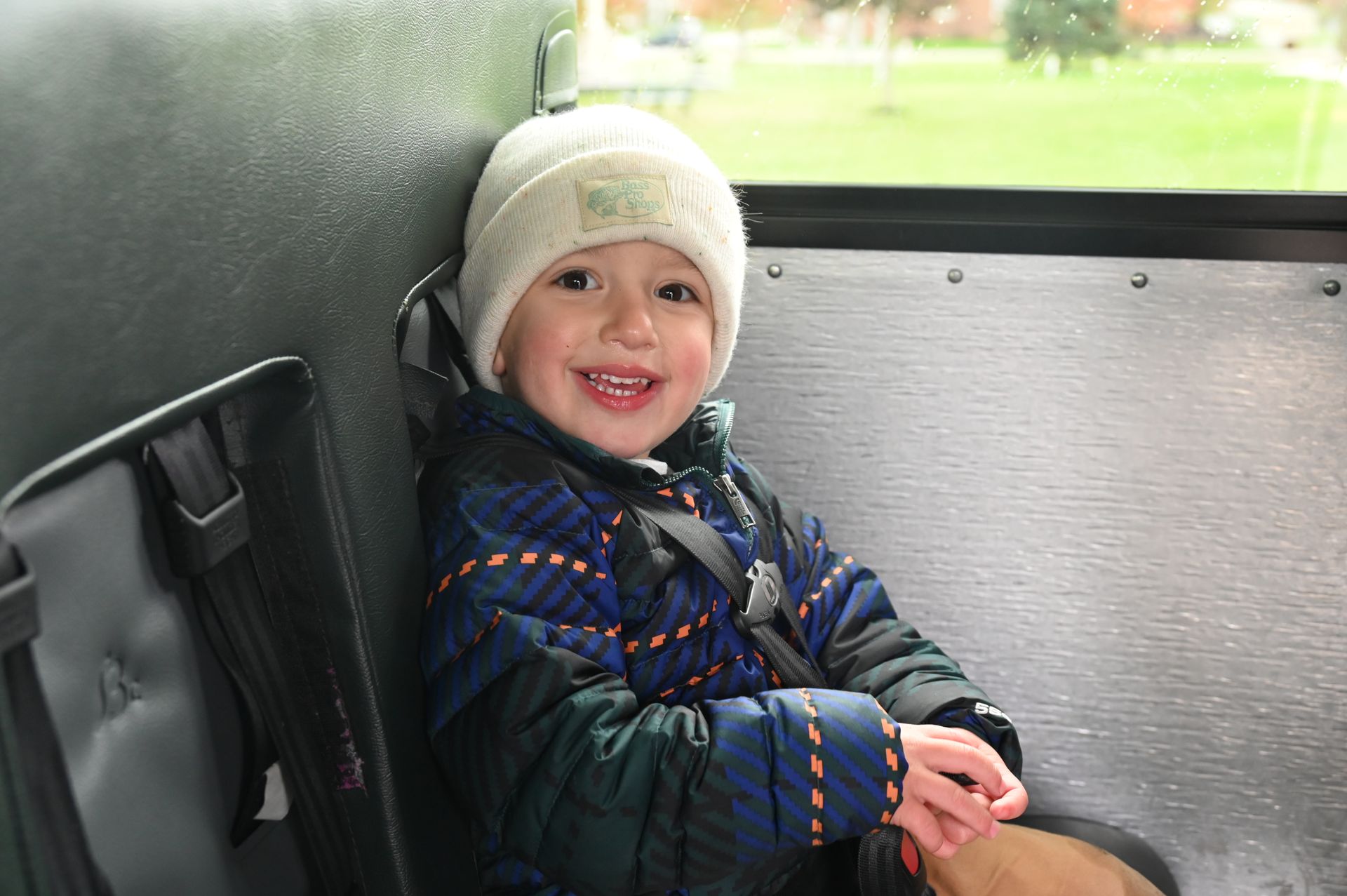 Child in winter coat and hat smiles on a bus, secured by a seatbelt.