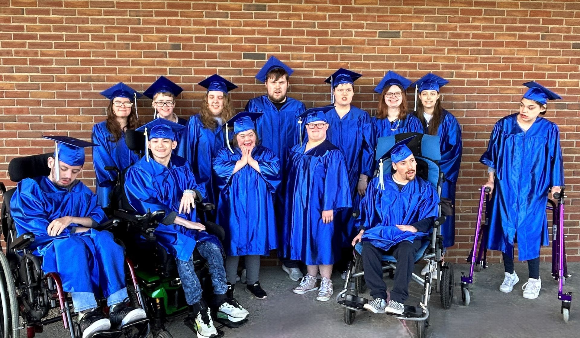 Group of people in blue graduation gowns and caps posing in front of a brick wall. Some use wheelchairs or walkers.