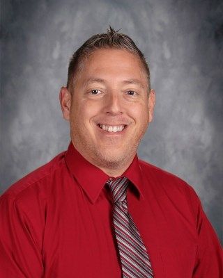 Man smiling, wearing a red shirt and striped tie, against a gray background.