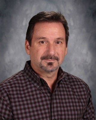 Man with short dark hair, mustache and goatee, wearing a patterned button-down shirt, looking forward.