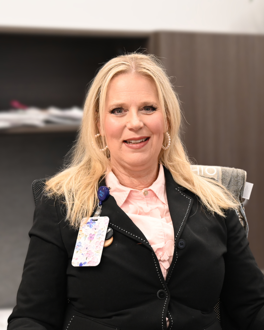 Woman with blonde hair, wearing a patterned top and necklace, smiling.