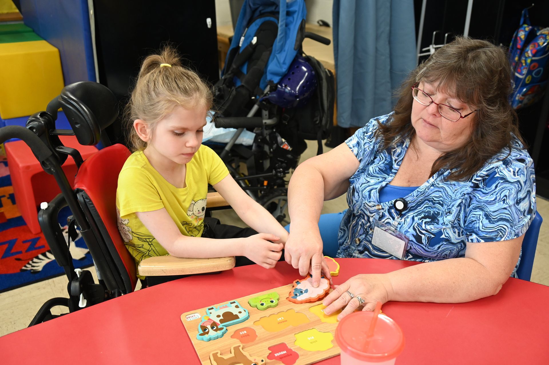 Girl in wheelchair doing puzzle with an adult, at a red table.
