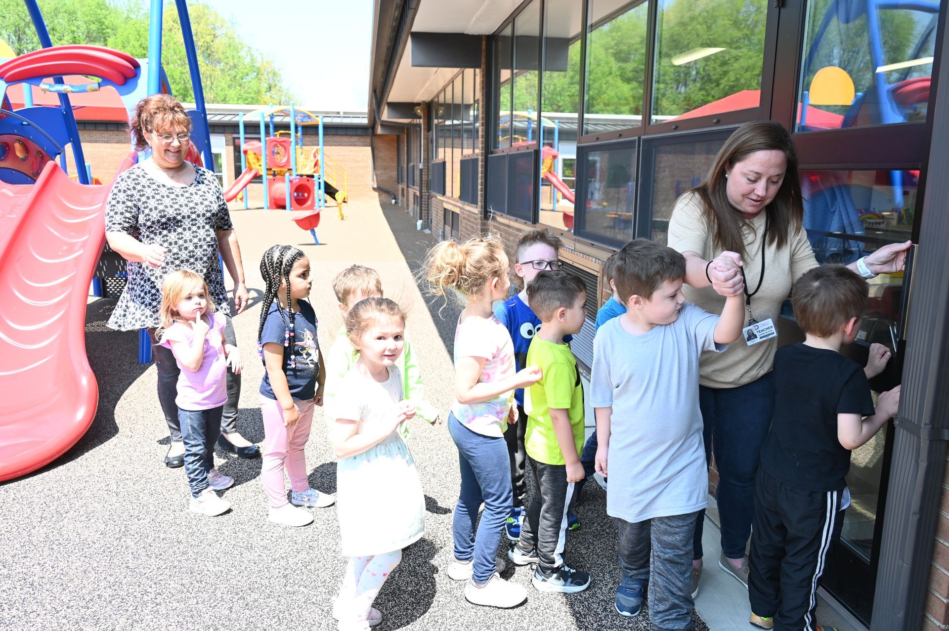 Children line up with teachers at a door to enter a building with a playground nearby.