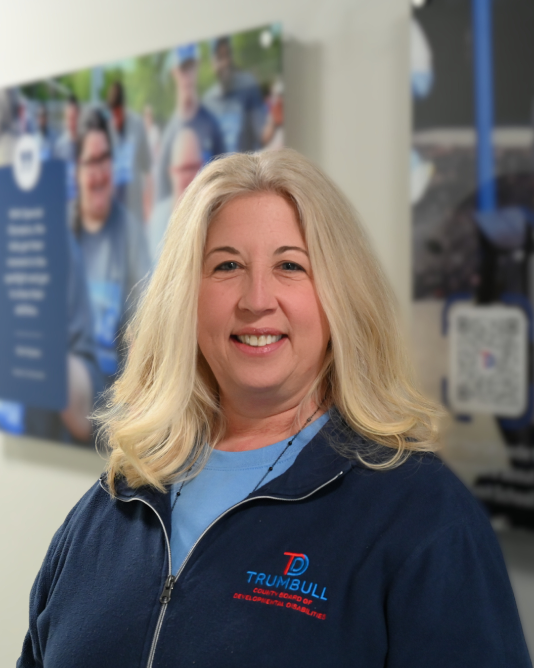 Woman with long blonde hair, smiling, wearing a teal blouse, against a blurred background.