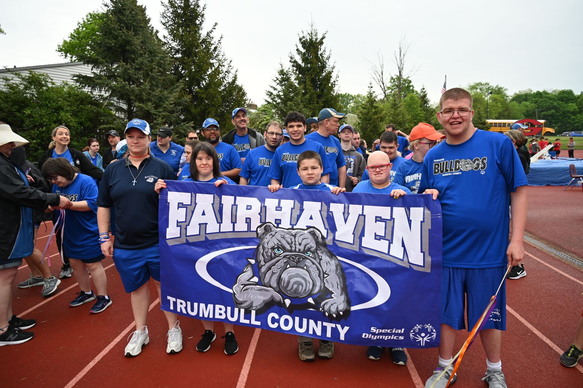 Fairhaven team with banner at a track. Blue and white colors, smiling, outdoors.