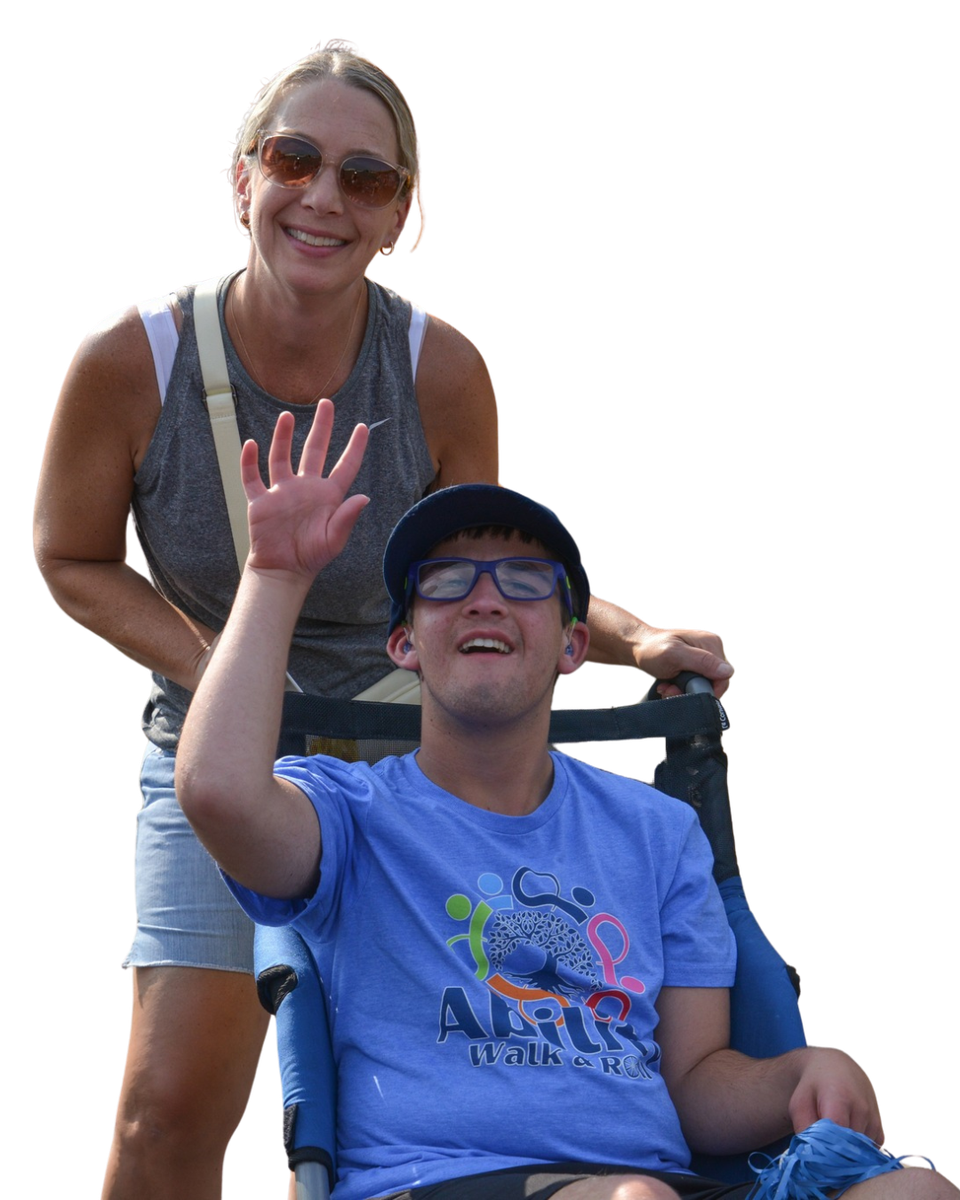 Woman pushing a wheelchair with a person waving. Both wear glasses and are smiling. Outdoor setting.