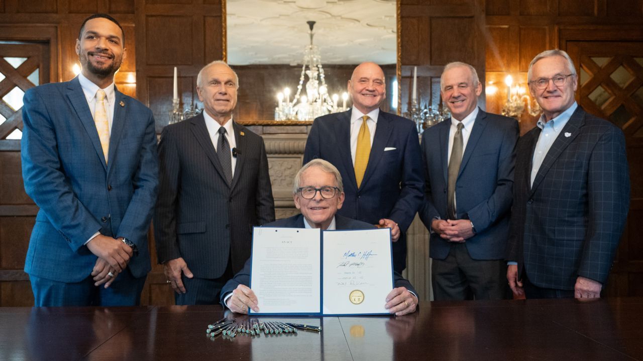 Group of men in suits surrounding Mike DeWine holding a signed document. Ornate room setting.