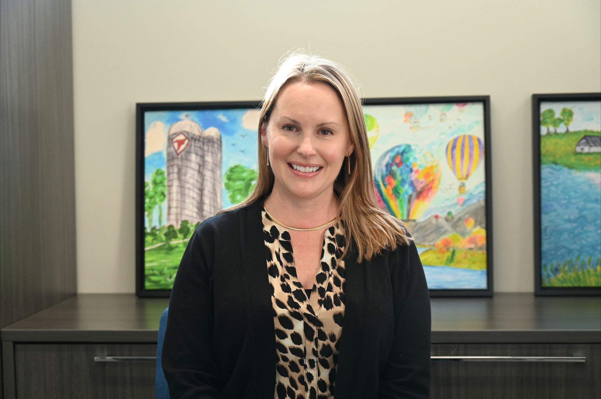 Woman with long blonde hair, smiling, wearing a necklace. Dark blue background.