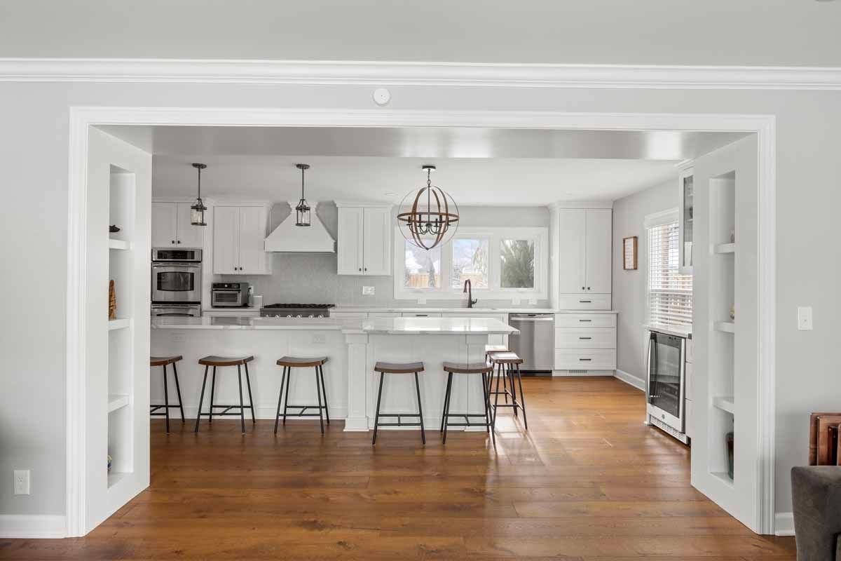 Bright kitchen with white cabinets, island with stools, and wooden floors. Seen through doorway.