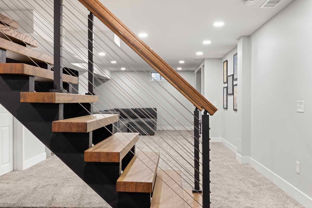 Wooden staircase with black supports and cable railings leading down to a finished basement.