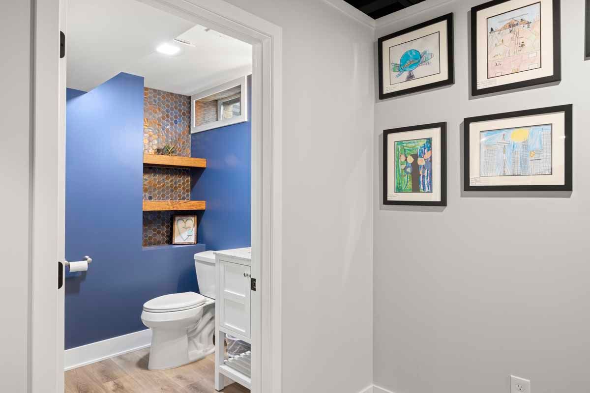 Blue bathroom with shelves, toilet, and framed artwork. Gray walls and black ceiling.