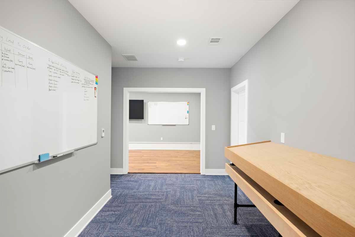 Hallway with blue carpet, whiteboard, and wooden desk. Another room has a whiteboard and black screen.