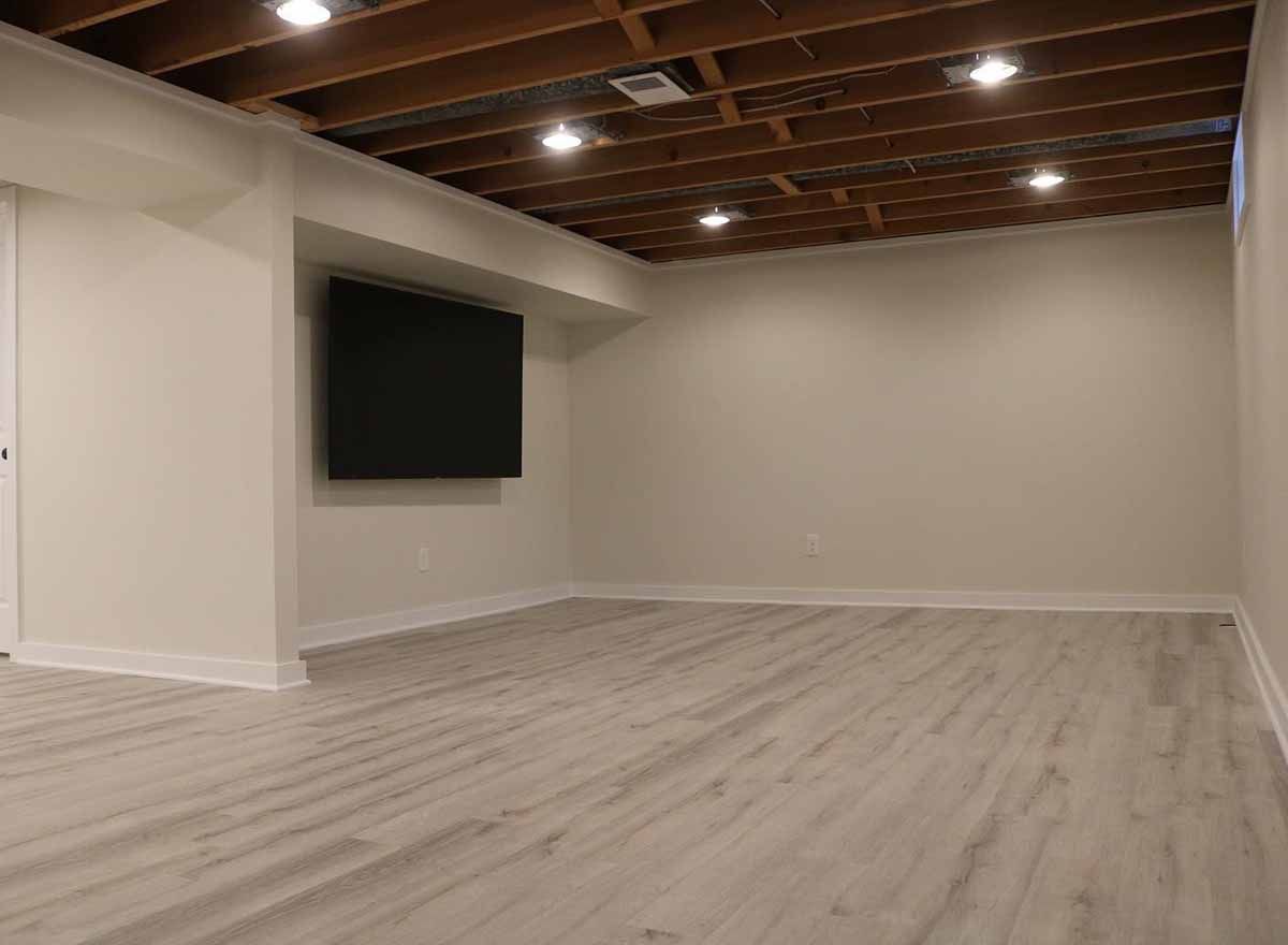 Empty, bright basement room with wood-look flooring, white walls, recessed lighting, and a wall-mounted TV.