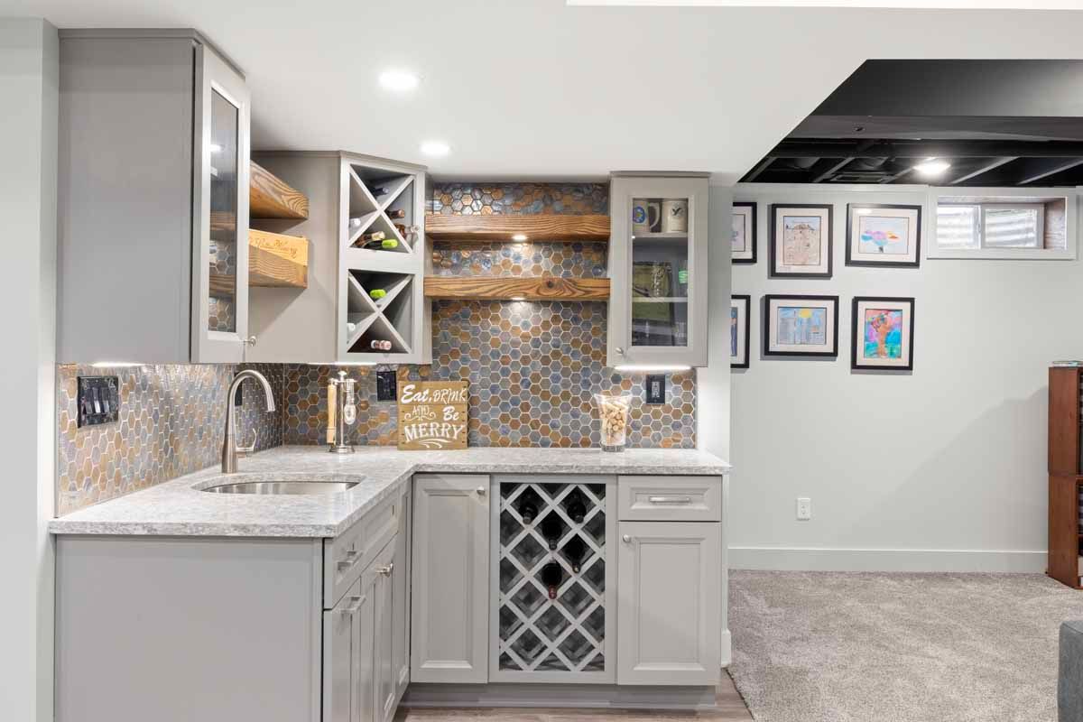 Gray basement bar with cabinets, sink, wine rack, and colorful backsplash; framed art on the wall.