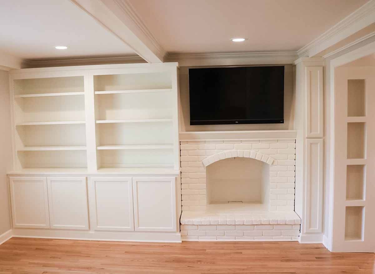 Built-in white bookshelves and cabinets flank a white brick fireplace with a TV mounted above.