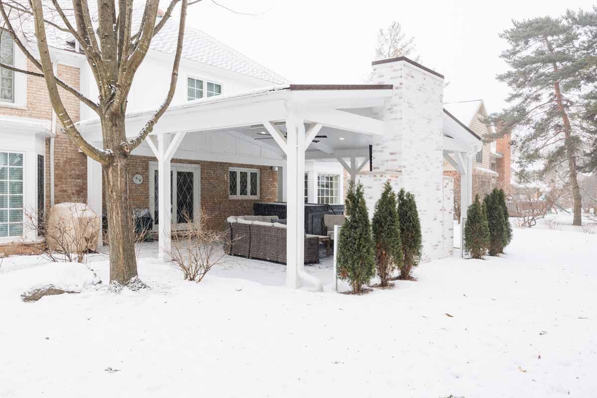 Snowy backyard with a white pergola and outdoor kitchen, trees, and a brick house.