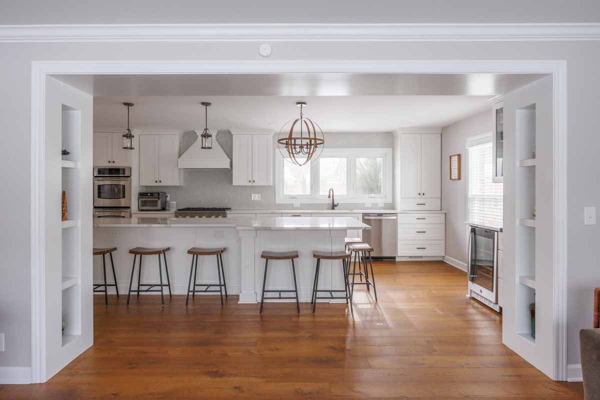 Kitchen interior with white cabinets, island with stools, and a chandelier; viewed through doorway.
