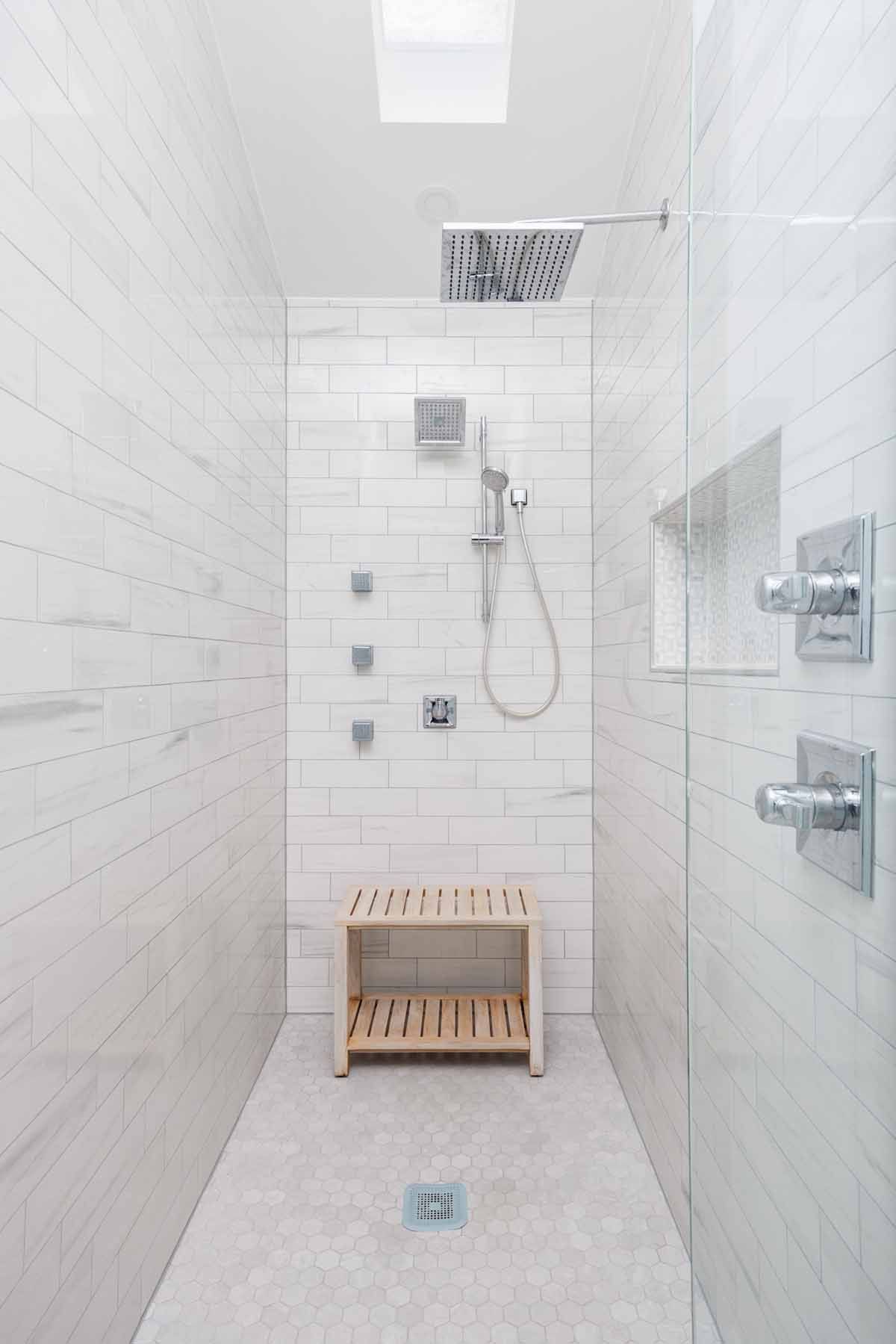 White tiled shower with glass wall, rain shower head, bench, and multiple body sprayers.