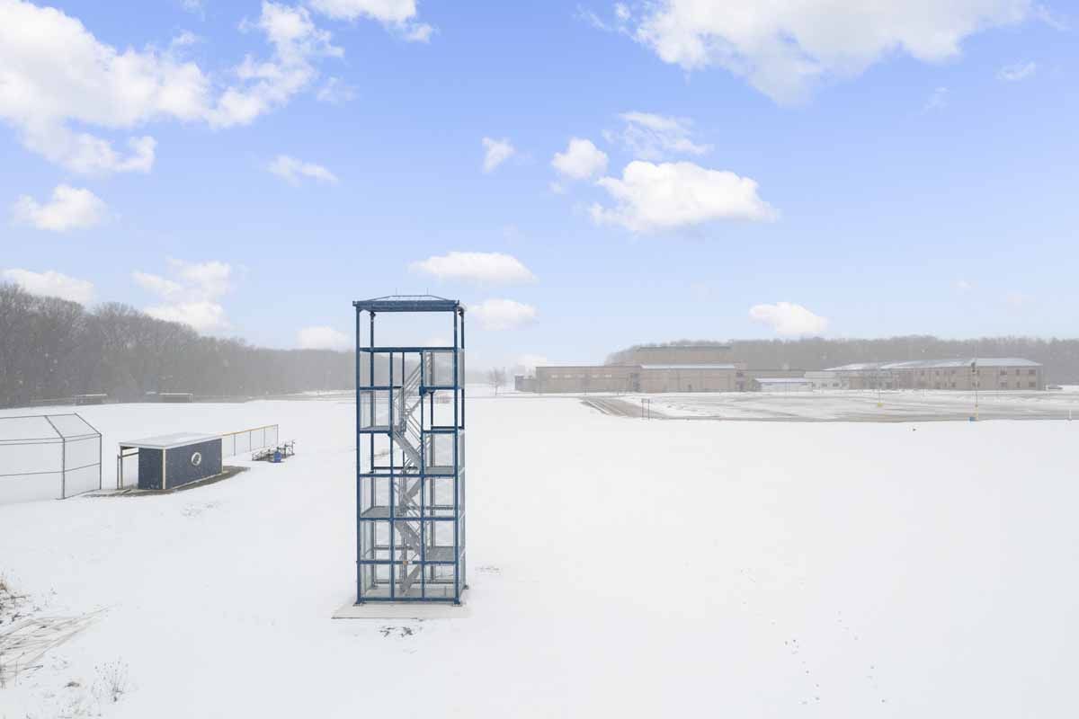 Snowy outdoor scene with a blue metal tower, buildings in the background, and a partly cloudy sky.