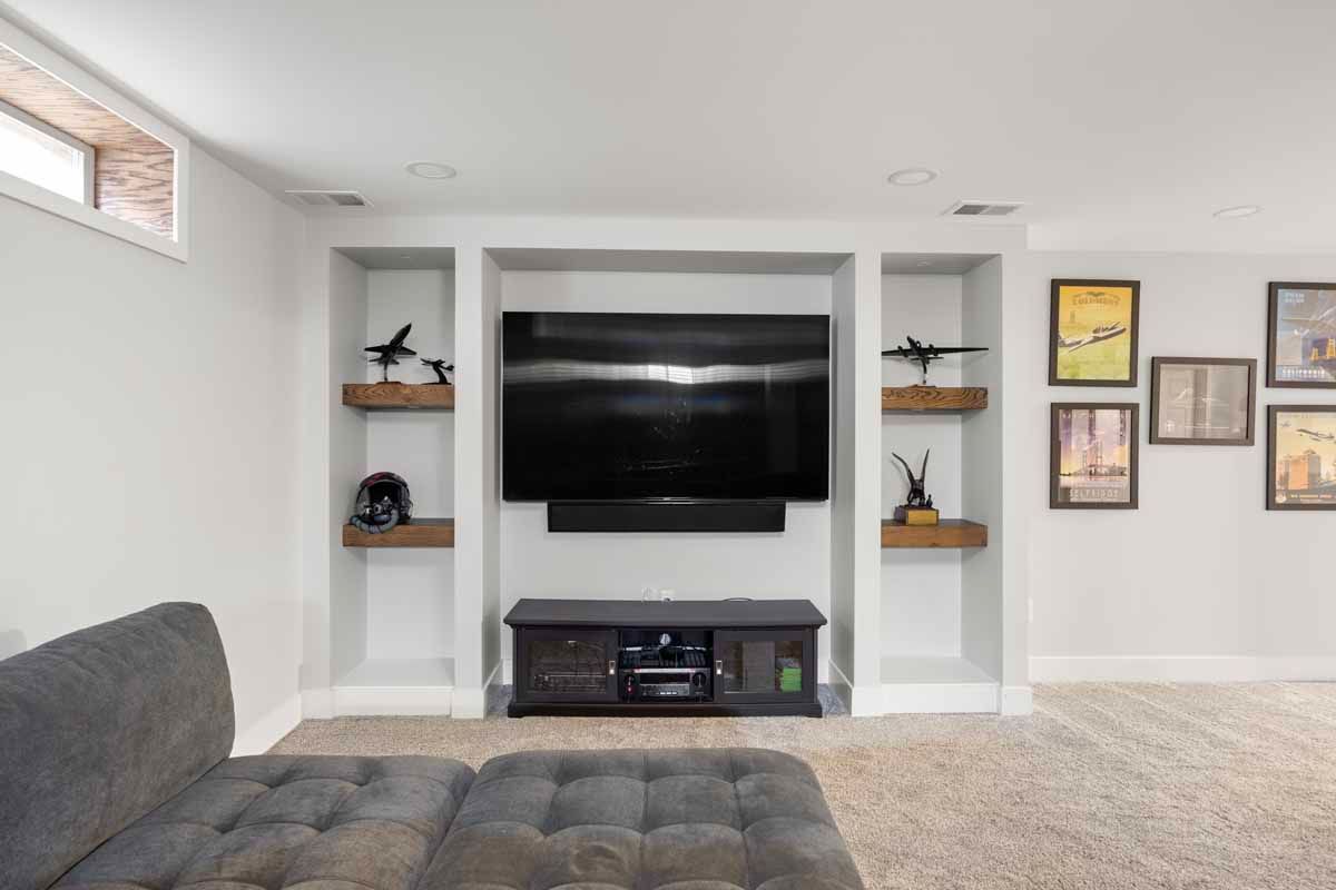 Built-in media center with a large TV, sound bar, and shelving. Gray sofa, tan carpet, and framed art.