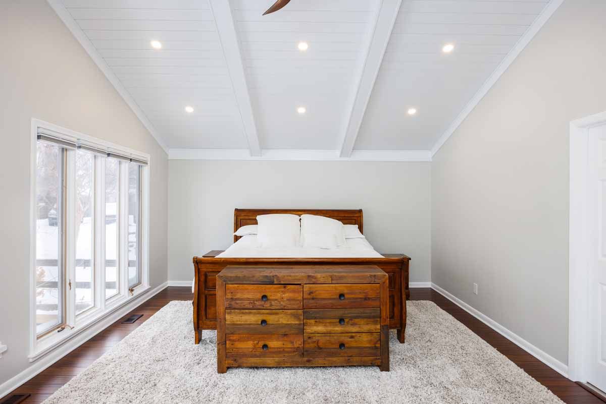 Bedroom with a wooden bed, dresser, and white rug. White walls and a sloped ceiling with recessed lights.