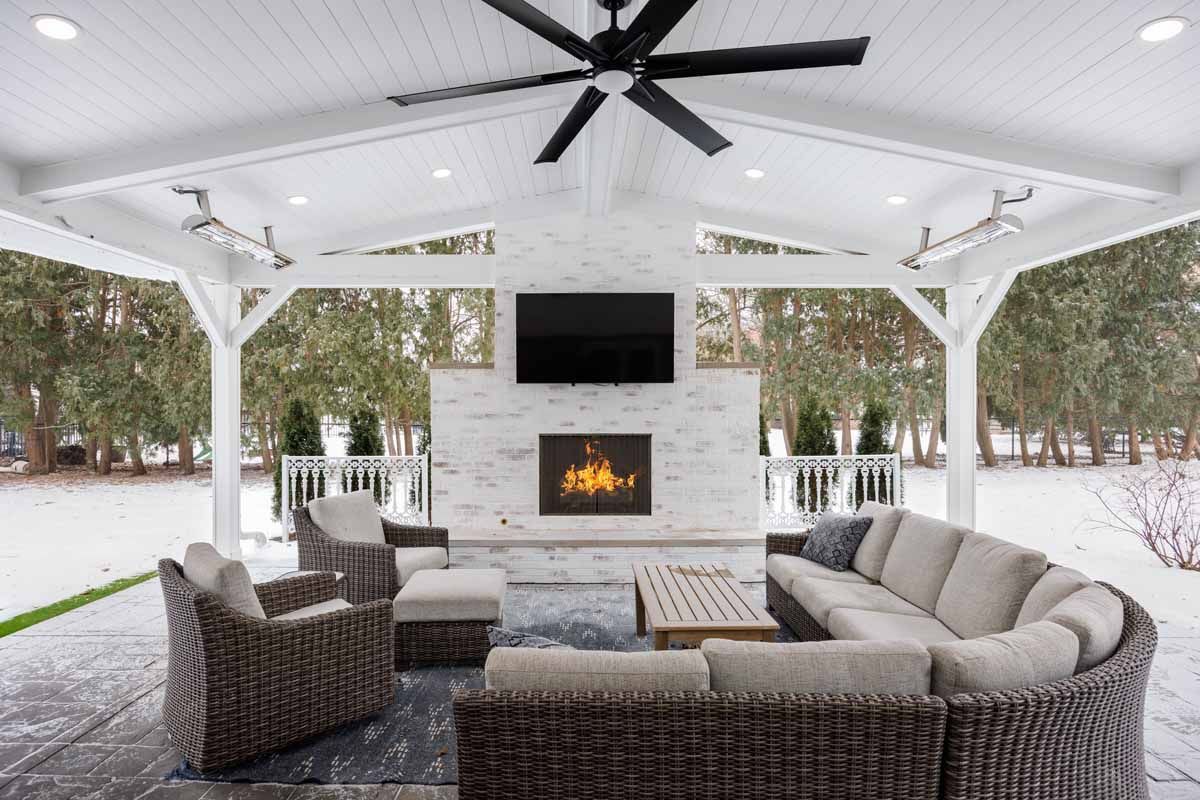Outdoor patio with fireplace, TV, and seating under a white-painted, gabled roof; winter landscape in background.