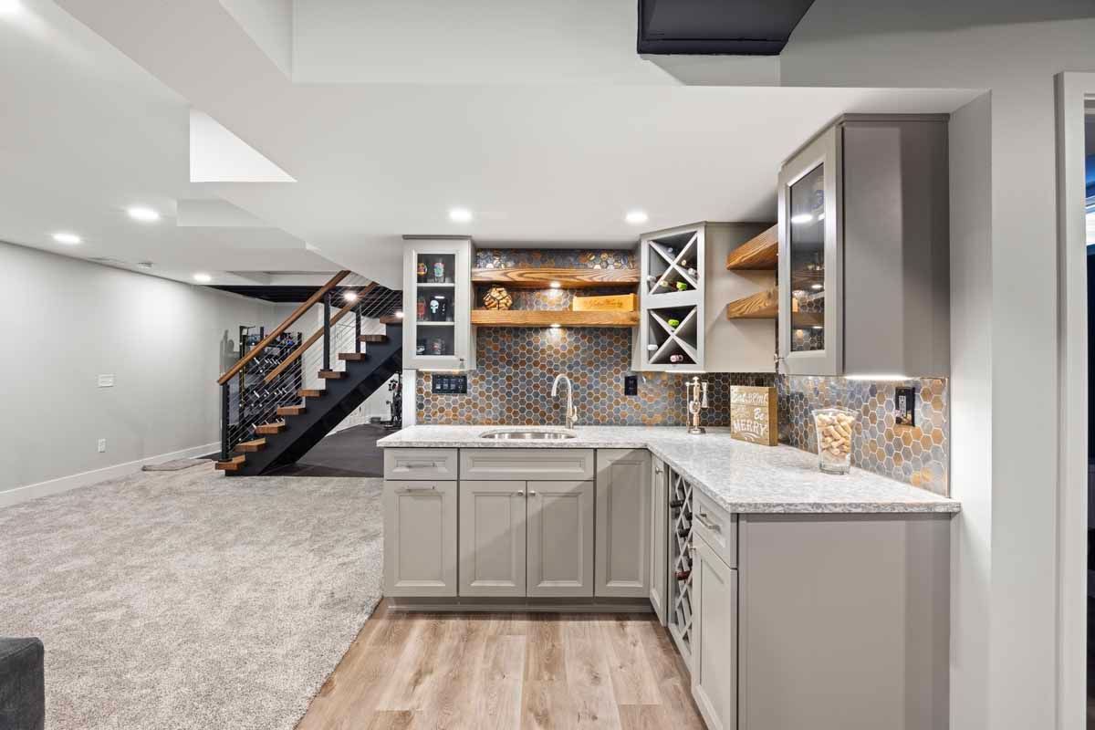 Gray basement with a kitchen area, wooden stairs, and a built-in bar with grey cabinets and a sink.