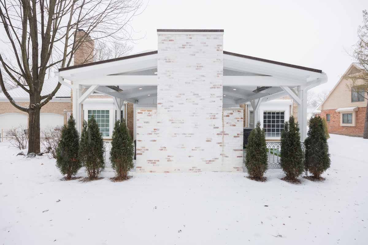 A snow-covered white brick outdoor structure with evergreens, two windows, and a chimney.