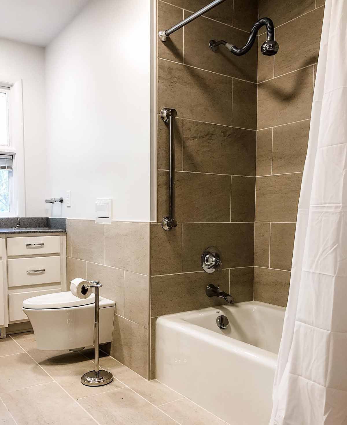 Accessible bathroom with a tub, shower, toilet, and grab bar. Neutral tiles and a white shower curtain.