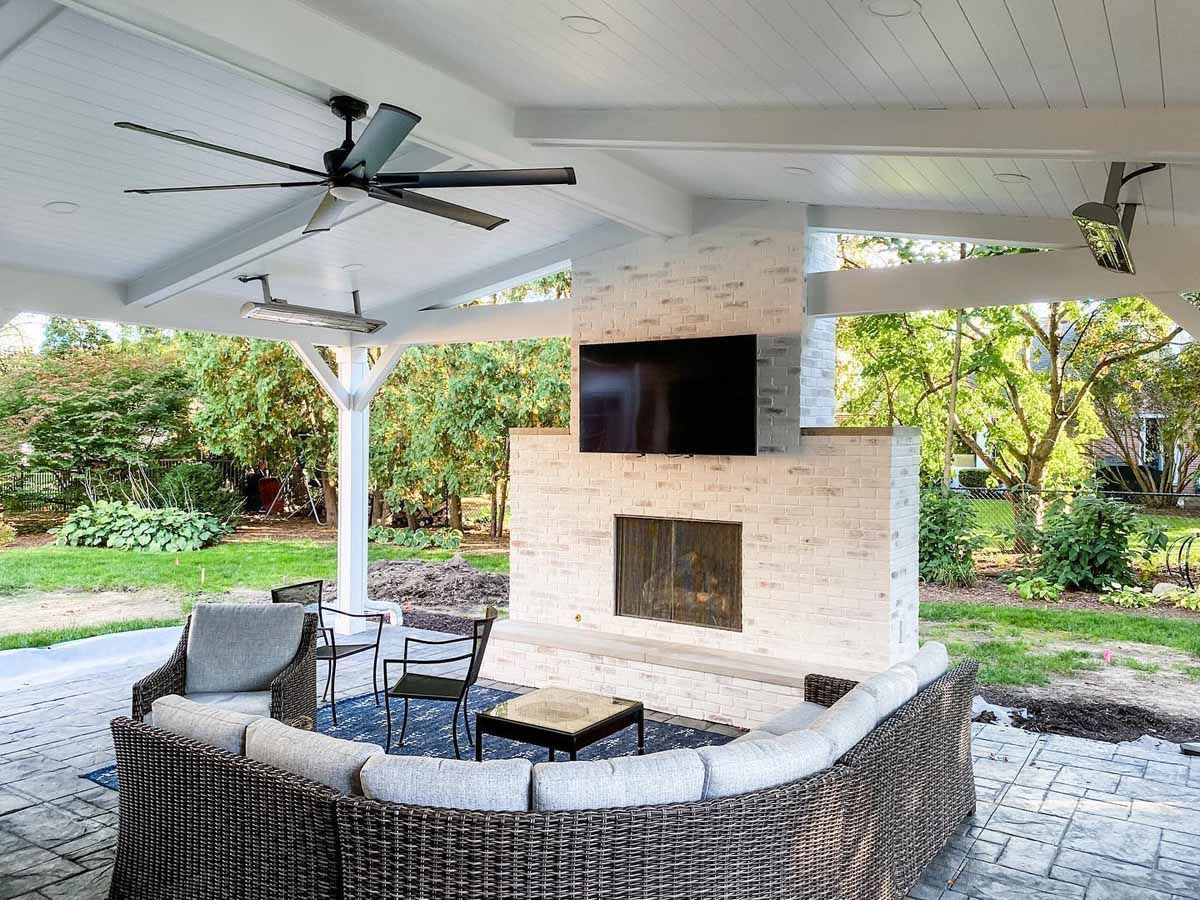 Outdoor patio with brick fireplace, TV, fan, and wicker seating under a white pergola.