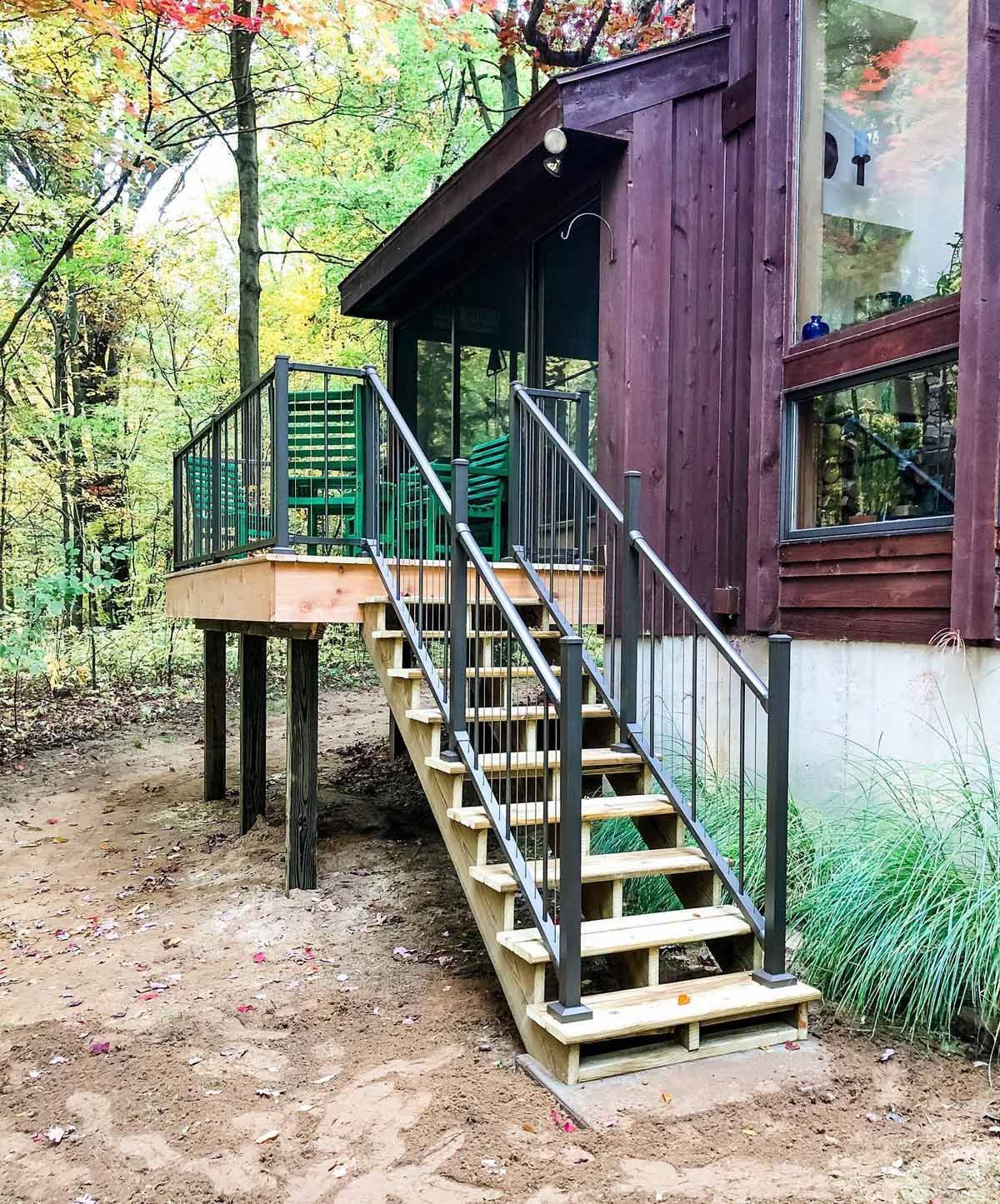 Wooden deck and stairs with black railings leading to a cabin in a wooded area.