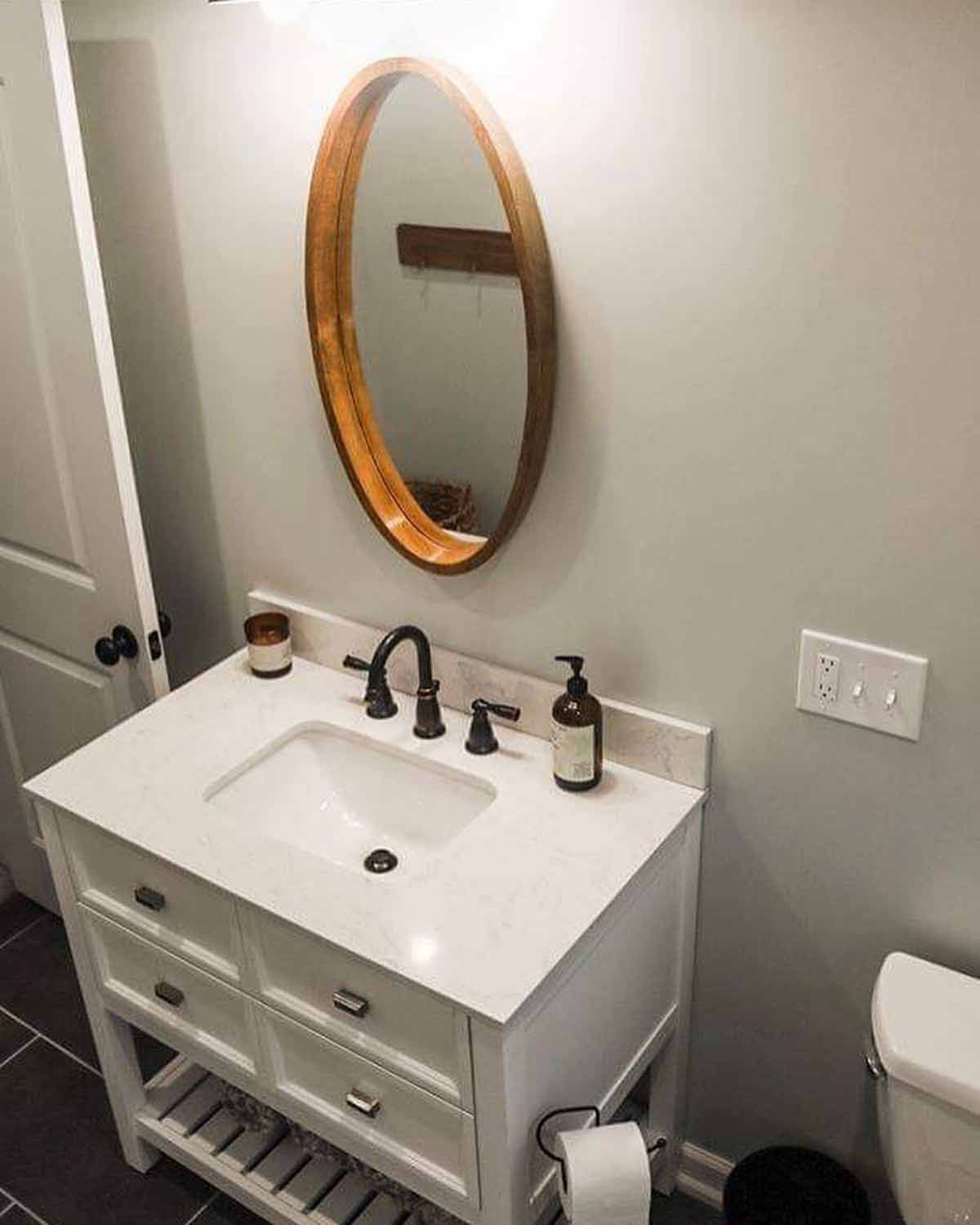 White bathroom vanity with oval wood-framed mirror and dark fixtures. Gray walls and floor, white door.