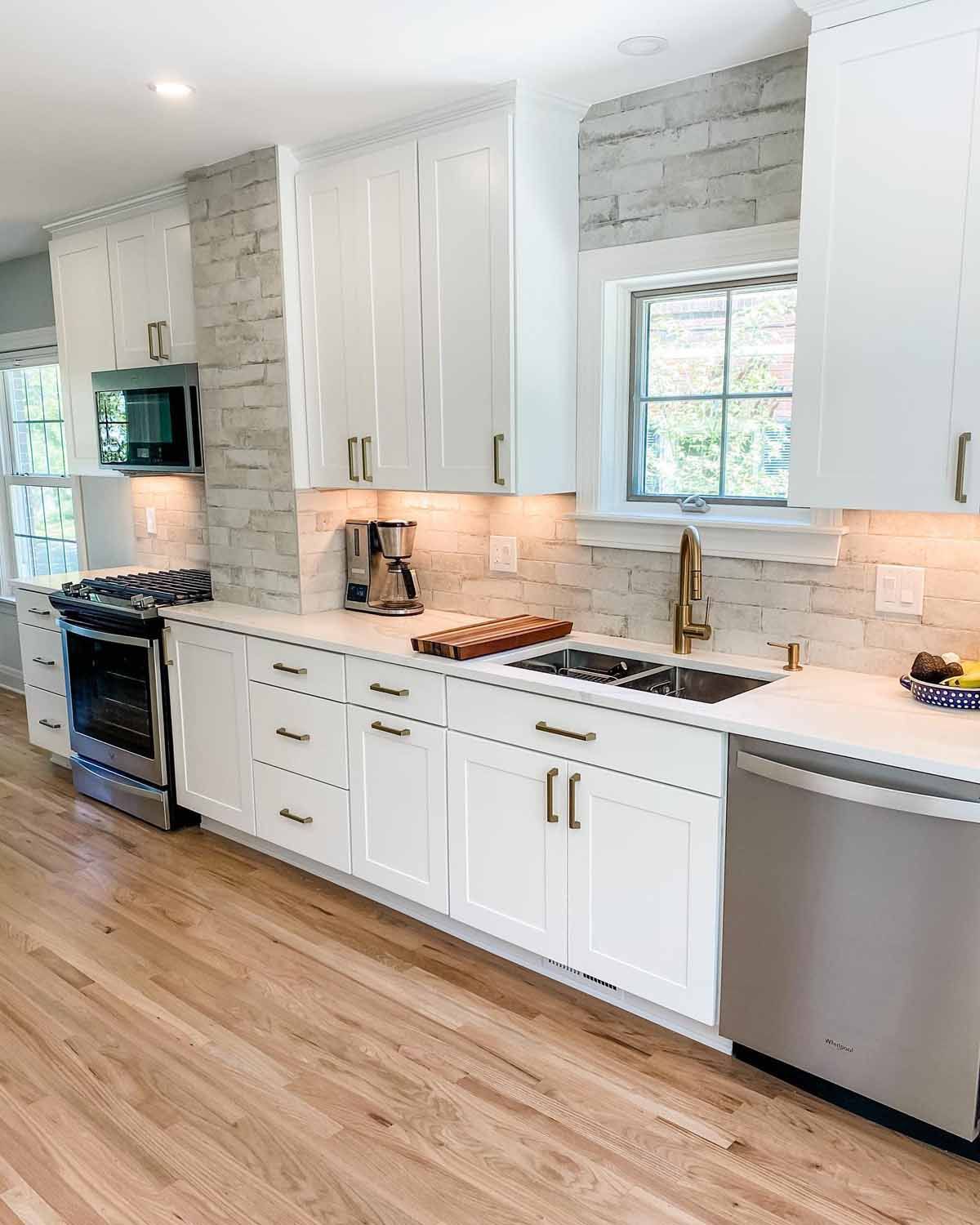 White kitchen with light wood floors, white cabinets, stainless steel appliances, and a brick-like backsplash.