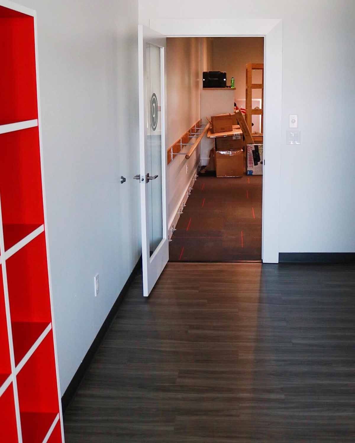 Open door leading to a room with boxes. Red shelving on the left. Wooden floor.