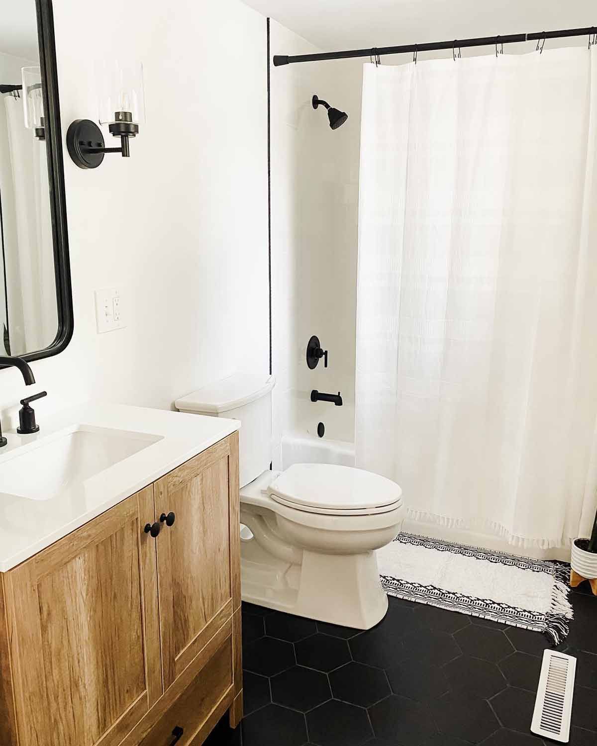 Bathroom with white walls, wooden vanity, black hexagonal floor, and white shower curtain.