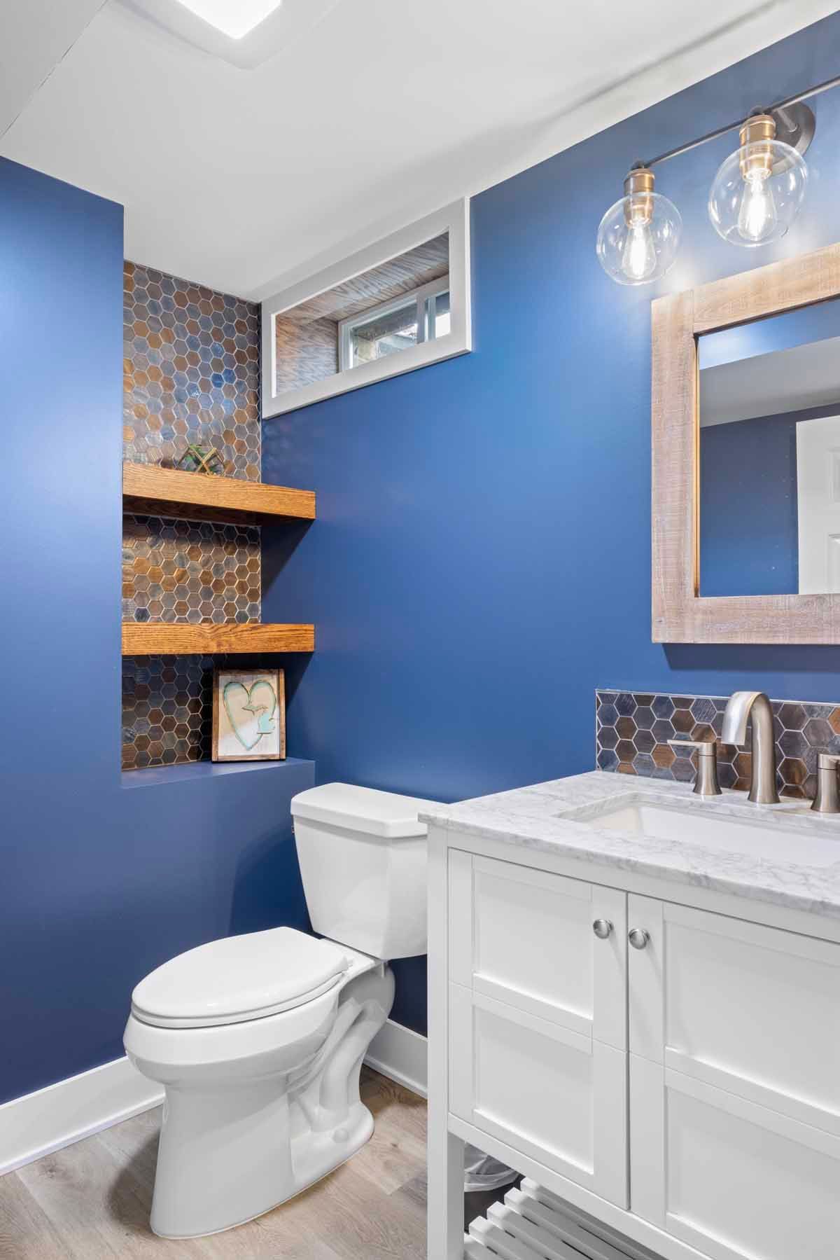 Blue bathroom with white toilet, vanity, and mosaic tile accent wall, with floating shelves.